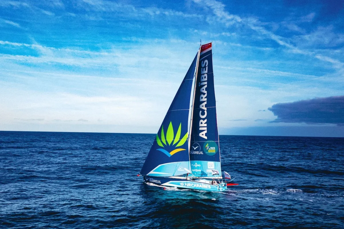 L'IMOCA Groupe Dubreuil II navigue au près sur l'océan, sa grand-voile et son foc arborant les logos Air Caraïbes et Groupe Dubreuil sous un ciel bleu.